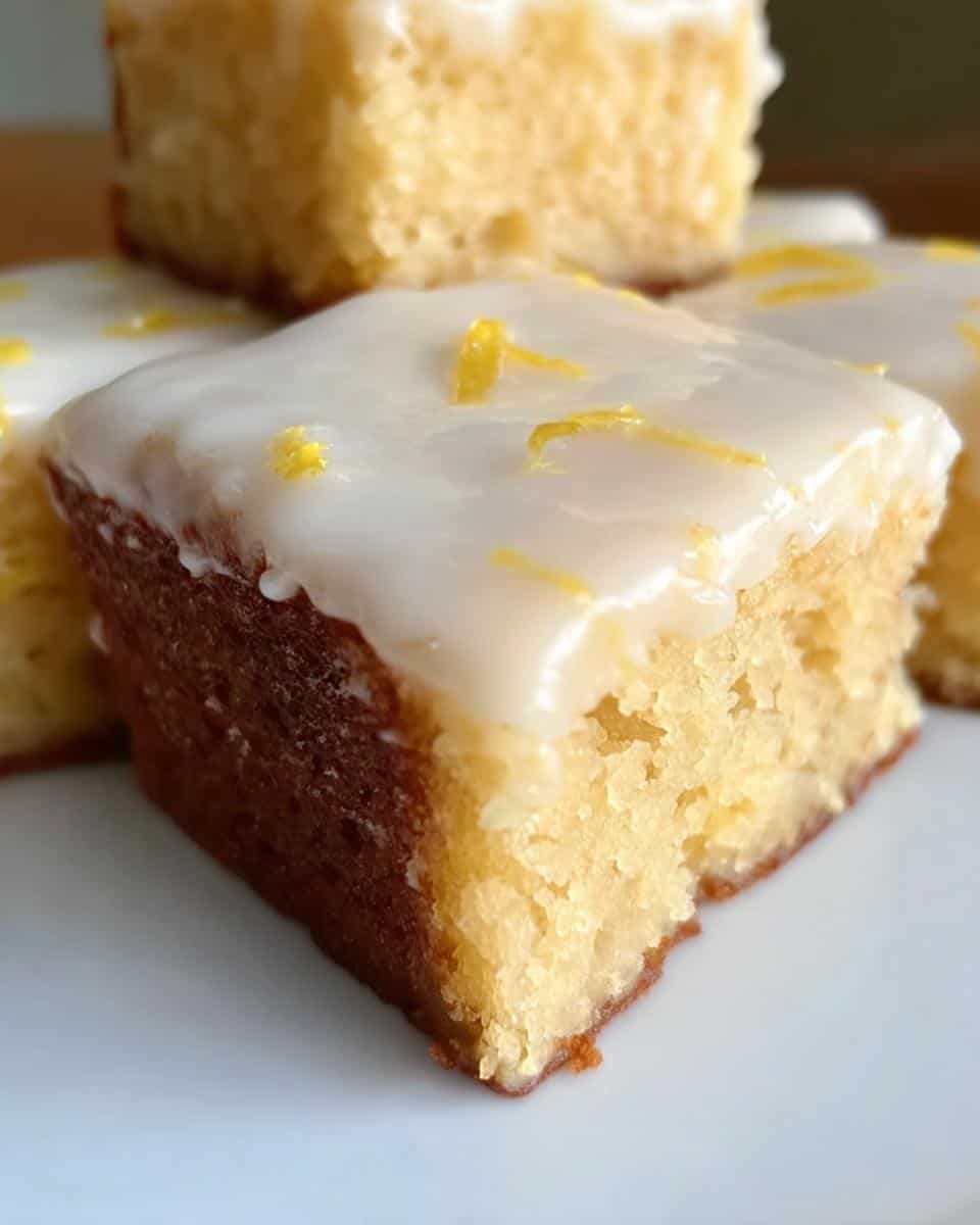 Fluffy lemon cake square with white icing and lemon zest topping, close-up.