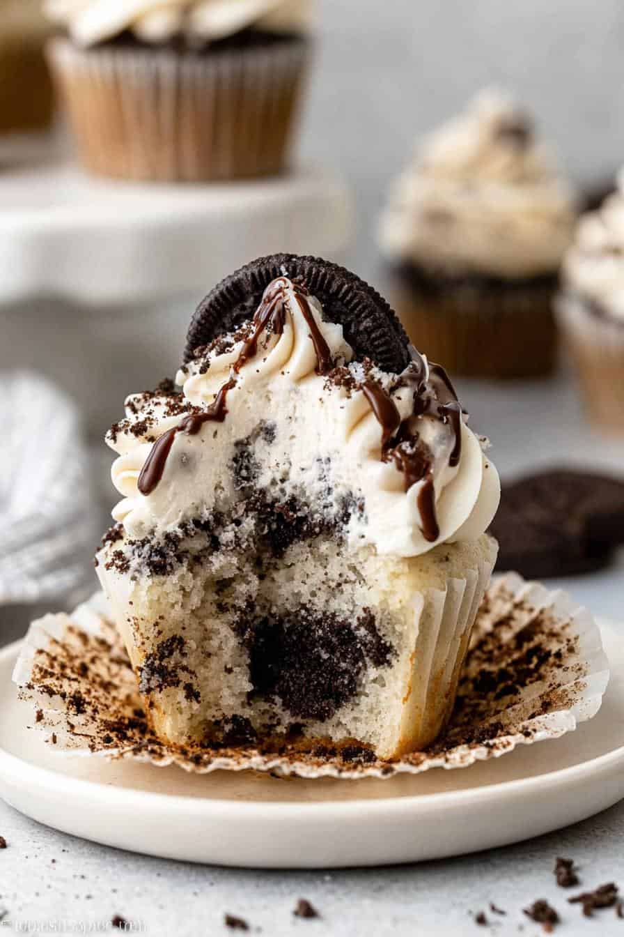 Oreo cookie cupcake with creamy frosting, chocolate drizzle, and crushed cookies, served on a white plate with a rustic background.
