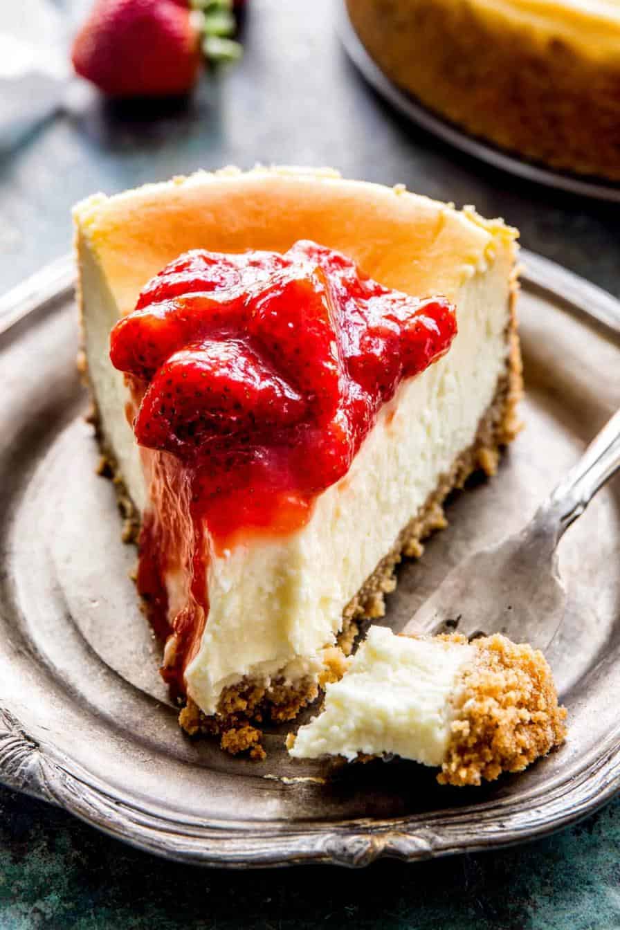 Rich strawberry cheesecake with graham cracker crust on silver tray, close-up, with a fork and fresh strawberries in background.