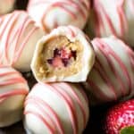 Strawberry-filled vanilla chocolate-covered cake balls decorated with pink drizzle, fresh strawberries, and a textured dark background. Perfect treat for celebrations and desserts.