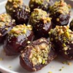 Rich chocolate-covered date treats topped with chopped pistachios and crunchy cookie crumbs, perfect for healthy snacking and satisfying sweet cravings. Freshly prepared and beautifully presented on a white plate.