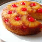 Fresh pineapple upside-down cake with cherries on top, served on a white plate. Delicious homemade dessert featuring caramelized fruit and moist cake layers. Perfect for a sweet treat or special occasion.