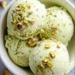 Creamy pistachio ice cream scoops topped with chopped pistachios in a white bowl on gray background, delicious homemade frozen dessert, pistachio-flavored ice cream, pistachio nuts, ice cream recipe, dessert ideas, The Bakers Whisk.