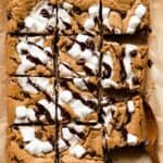 Rich salted caramel gluten-free cookie bars topped with marshmallows and chocolate drizzle on parchment paper. Ideal for decadent dessert recipes and cookie bar favorites.