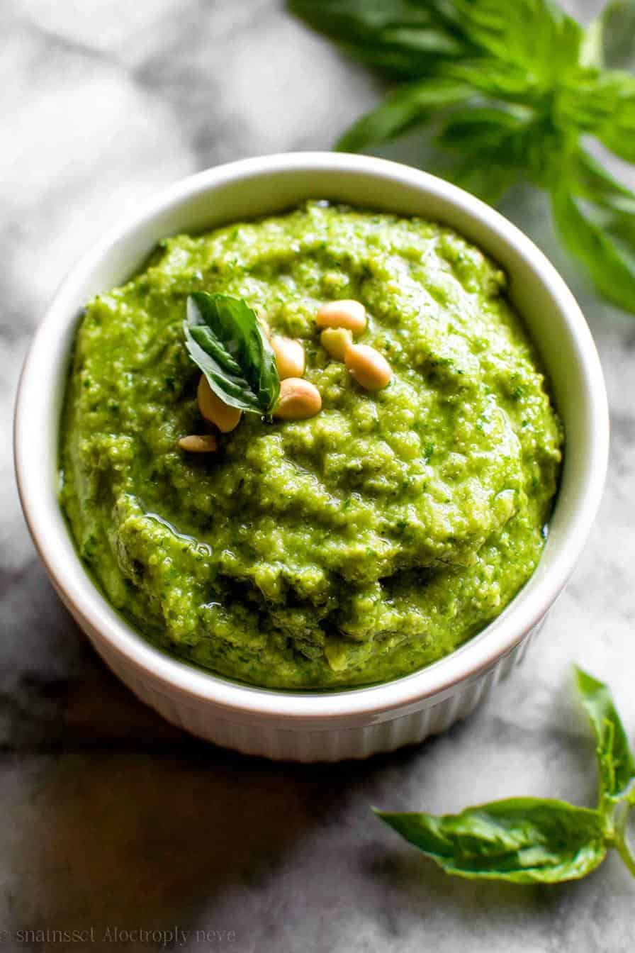Creamy basil pesto dip with pine nuts and fresh basil garnish in a white ramekin on a textured gray surface.